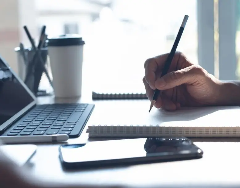 A person's hand is seen taking notes in front of a computer, with a phone on the table and a coffee cup in the background